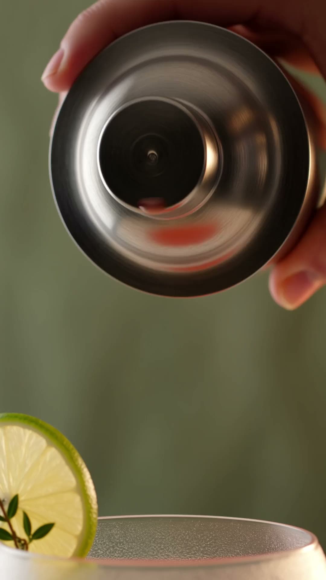 A coral cocktail poured from a steel shaker into a coupe with a lime wheel and thyme.
