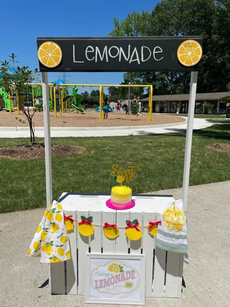The Child Stand dressed as a lemonade stand, with a chalkboard Lemonade sign, lemon garland and a yellow cake, in a park.