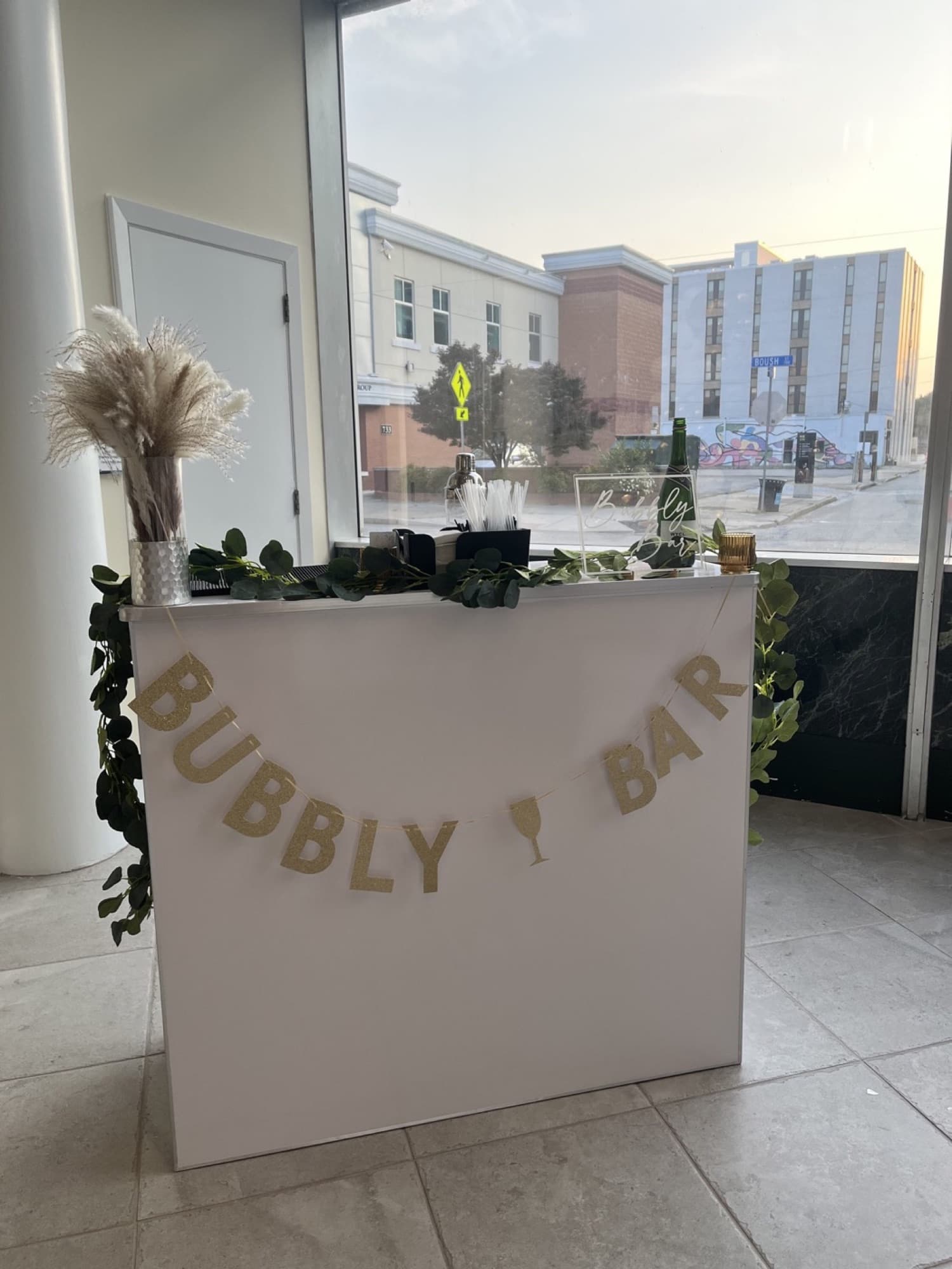 The Pop Up Bar dressed as a Bubbly Bar, with gold letters, pampas grass and greenery, by a venue window.