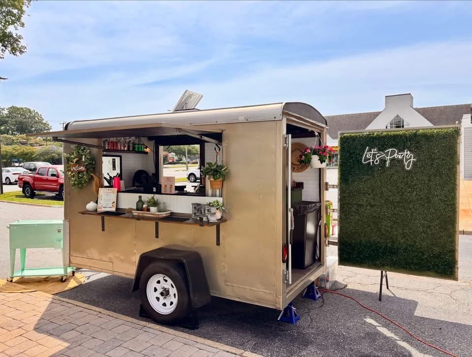 The trailer with its awning up and the Let's Party greenery wall beside it, on a sunny lot.